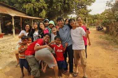 Volunteers and local children crowded together on a dirt road, all pulling faces for the camera