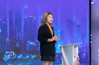 Nhi standing at the candidate's podium on the Cơ hội cho ai set, against a blue city backdrop